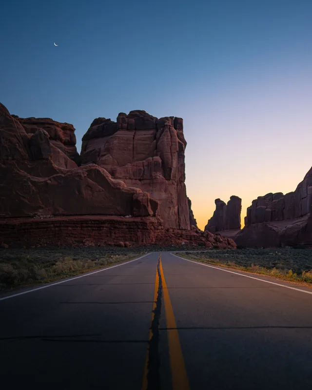 A desert road running between red sandstone towers under a crescent moon