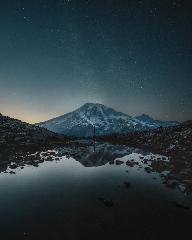 A figure with a headlamp below a snow-covered peak under the stars